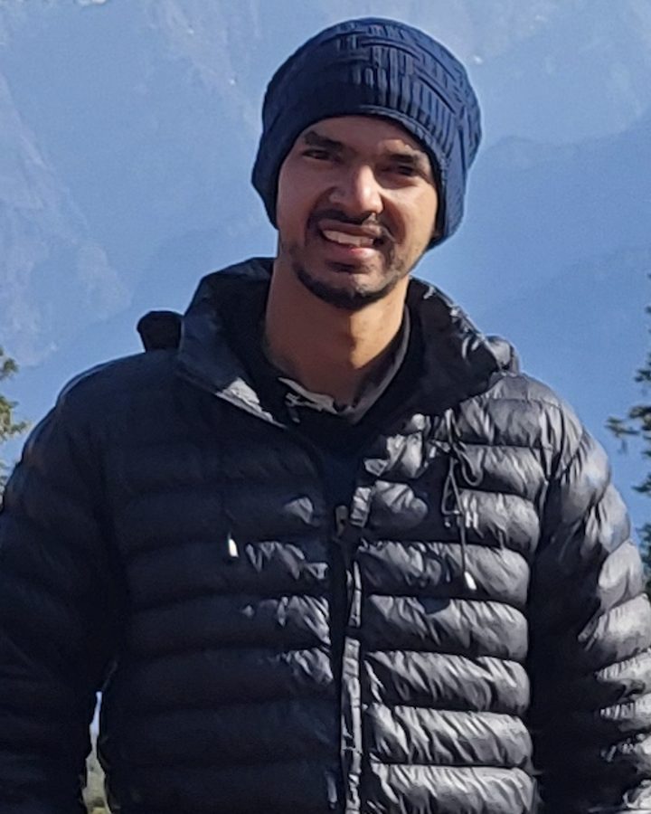 Charchit Sharma in a beanie and puffer jacket, mountains behind him